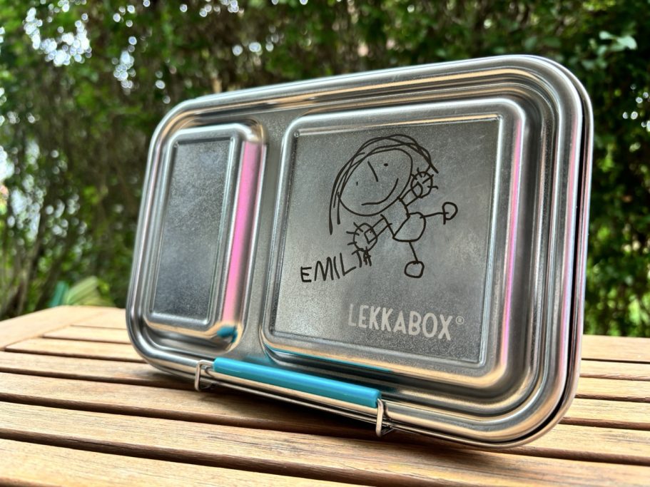 Edelstahl-Bentobox mit gezeichnetem Charakter und buntem Deckel, auf einem Holztisch.