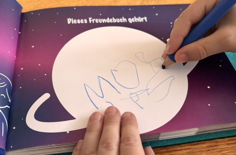 geöffnetes Freundebuch Weltraum. Kind schreibt mit einem blauen Stift seinen Namen in einen weißen Saturn.