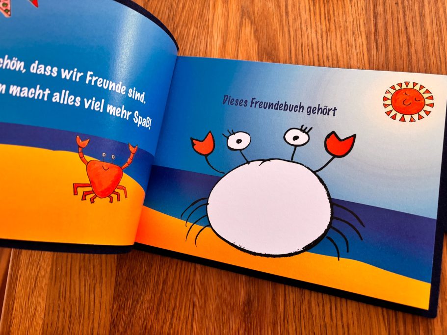 Ein offenes Kinderbuch mit bunten Illustrationen eines Krebses und einer Sonne am Strand.