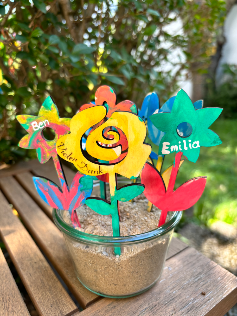 Bunte Papierblumen in einer Glasvase auf einem Holztisch im Garten.