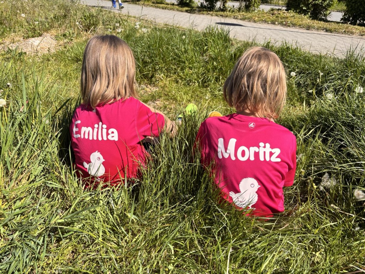 Zwei Kinder sitzen im Gras, tragen rote T-Shirts mit den Namen Emilia und Moritz.