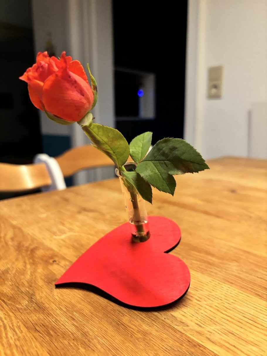 Eine rote Rose in einer herzförmigen Halterung auf einem Holztisch.